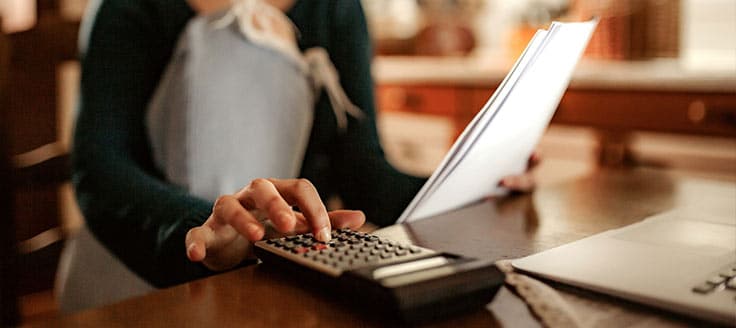 A woman uses a calculator as she refers to paperwork as she works with her invoicing software on her laptop.