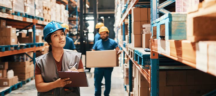 A warehouse worker takes inventory of stock on hand, using a pen and clipboard. Other workers walk in the background.