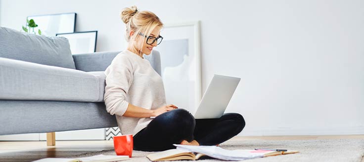 A woman works on her laptop while sitting on the floor of her living room.