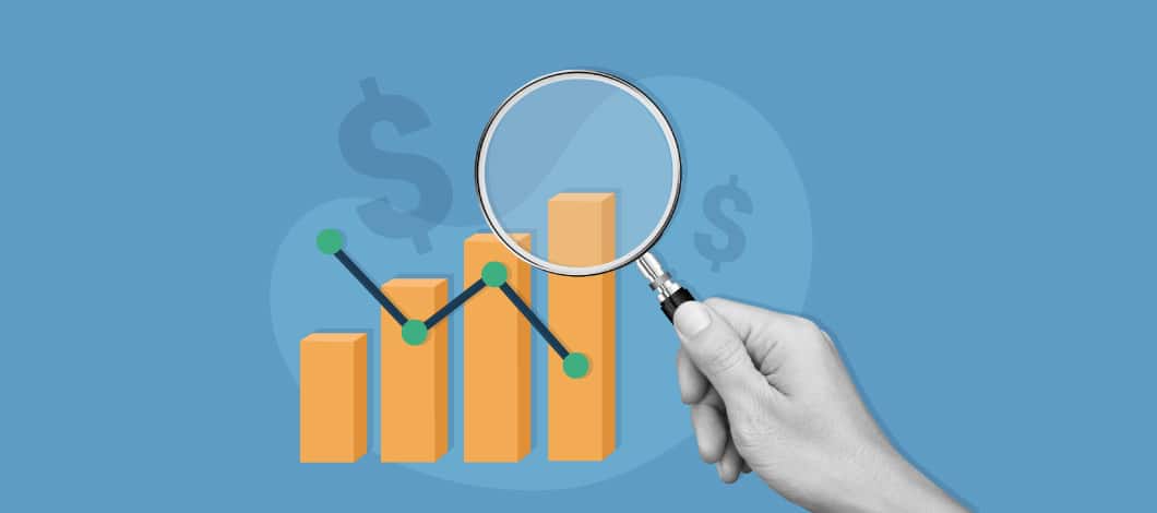 A hand holds a magnifying glass on a bar graph with dollar signs around